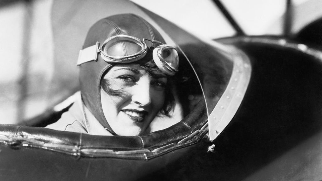 Black and white photo of historical woman wearing goggles in bi-plane.